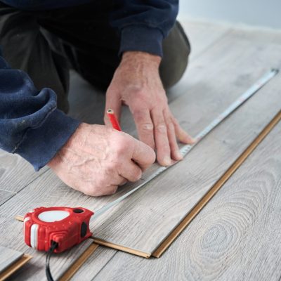 Close up of male hands using tape measure and pencil while drawing line on laminate plank. Man construction worker measuring laminate wooden board for floor installation. Home renovation concept.